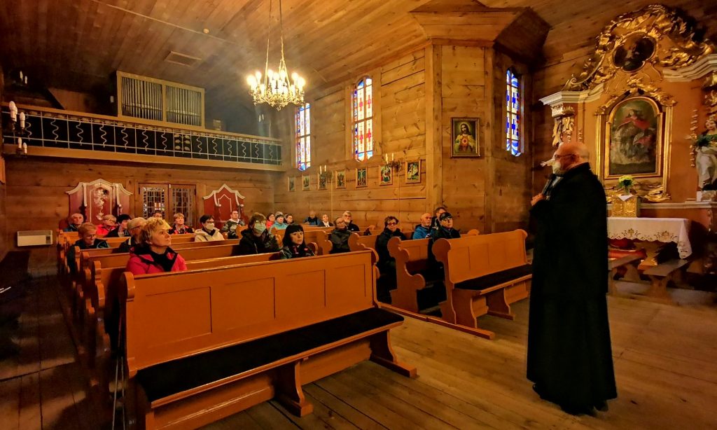 Wnętrze kościoła w Budziszewku, uczestnicy wycieczki siedzą w ławkach, ksiądz opowiada o kościele 