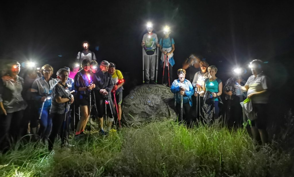 kilkanaście osób z kijkami i czołówkami stoi obok kamienia nocą